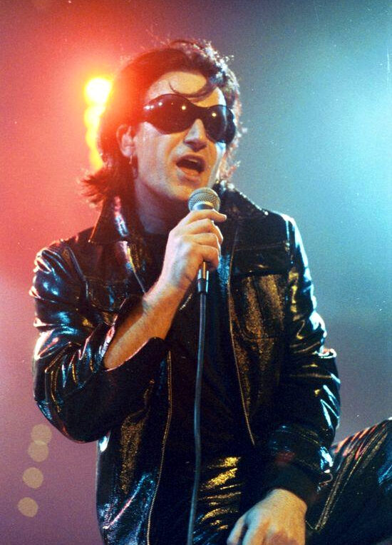 Bono of U2 in his The Fly persona on the 1992 Zoo TV tour, in sunglasses and leather, singing into a microphone